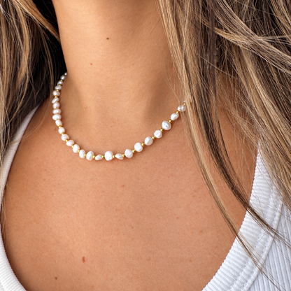 A close-up of a woman wearing a delicate strand necklace made of small white pearls with tiny gold spacer beads, styled with a white ribbed tank top.