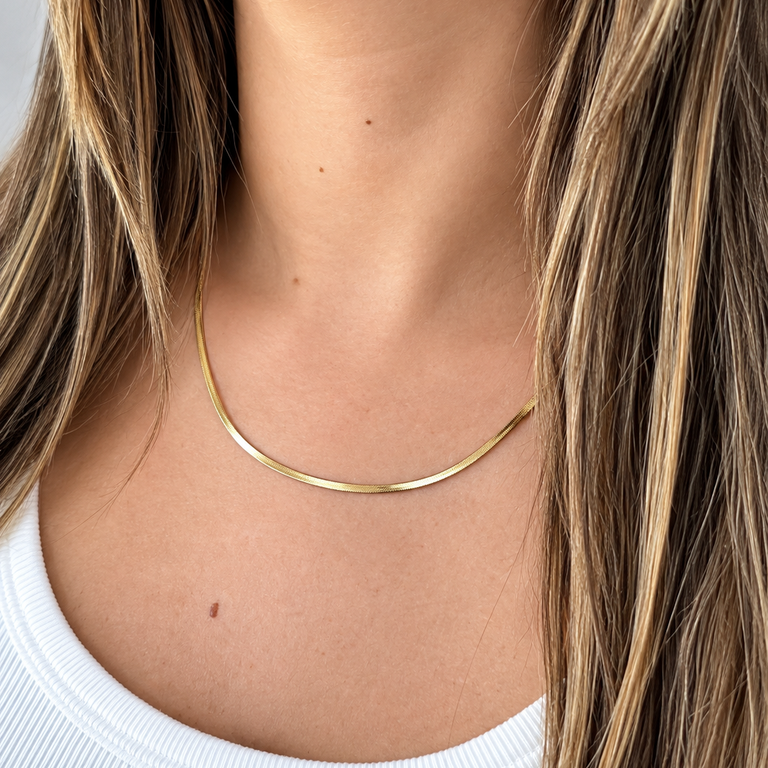 A close-up of a woman's neck and upper chest wearing a thin, polished gold chain necklace. She has long, highlighted brown hair and is wearing a white ribbed top.