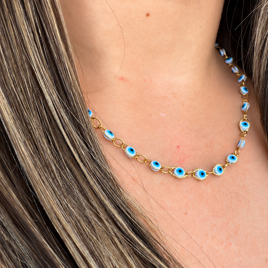 A close-up of a gold chain necklace with blue and white evil eye charms, worn on a model's neck with long brown hair.