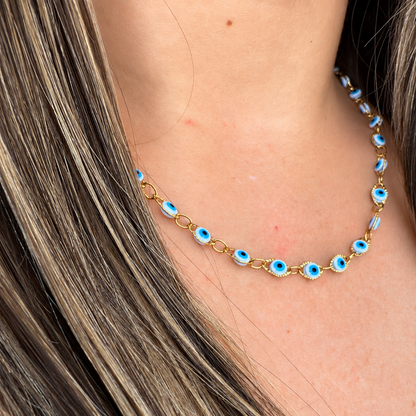 A close-up of a gold chain necklace with blue and white evil eye charms, worn on a model's neck with long brown hair.