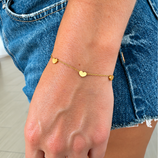 A delicate gold chain bracelet with three small heart charms worn on a person's wrist, paired with blue denim shorts.